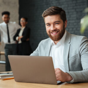 Image of cheerful young caucasian businessman sitting indoors using laptop computer. Looking camera.